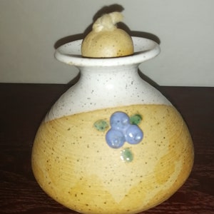 Op de afbeelding: Een kleine, witte en bruine keramische olielamp met een blauw en groen bosbessenontwerp. De lamp heeft een ronde, witte bovenkant met een lont.
