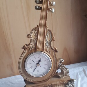 May include: A gold-toned, decorative clock shaped like a violin. The clock face has Roman numerals and a black hour and minute hand. The clock is on a white surface.