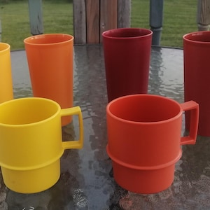 Peut inclure: Ensemble de six tasses et mugs en plastique colorés. L'ensemble comprend des gobelets hauts et cylindriques jaunes, oranges et rouges, ainsi que deux mugs jaunes et un orange avec des poignées. Les articles sont disposés sur une surface en verre.