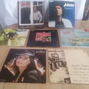 Może przedstawiać: Kolekcja starych płyt winylowych. Okładki przedstawiają portrety i teksty, z artystami takimi jak Adamo, Enrico Macias i Nana Mouskouri. Płyty są ułożone na drewnianej powierzchni, prezentując różne tytuły albumów.