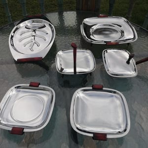 Op de afbeelding: Een set van zes roestvrijstalen serveerschalen met donkerbruine houten handvatten. De set bevat een grote ovale schaal, twee vierkante schalen, een vierkante schaal met een handvat en twee kleinere vierkante schalen.