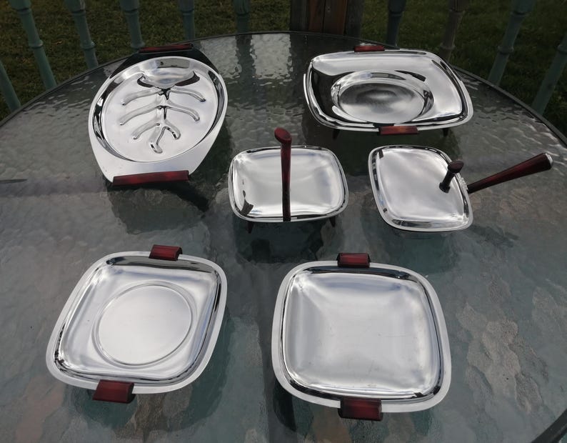 Op de afbeelding: Een set van zes roestvrijstalen serveerschalen met donkerbruine houten handvatten. De set bevat een grote ovale schaal, twee vierkante schalen, een vierkante schaal met een handvat en twee kleinere vierkante schalen.