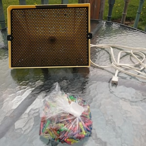 May include: A vintage Lite-Brite toy with a yellow frame and a black screen. A clear plastic bag filled with colorful pegs sits in front of the toy. A white power cord is connected to the toy.