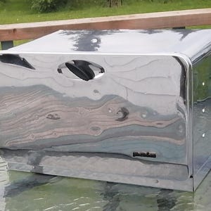 Pode incluir: Uma caixa de pão cromada brilhante, com cantos arredondados e uma pega na parte superior. Está colocada numa mesa com tampo de vidro, refletindo o ambiente circundante. A caixa tem uma forma retangular.