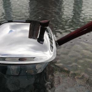 Op de afbeelding: Een glanzende, verchroomde steelpan met een donkerbruine houten handgreep. De vierkante pan reflecteert de lucht en de omgeving. De handgreep is ongeveer 20 cm lang en aan de zijkant bevestigd.