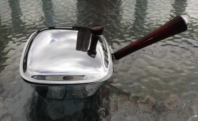 Op de afbeelding: Een glanzende, verchroomde steelpan met een donkerbruine houten handgreep. De vierkante pan reflecteert de lucht en de omgeving. De handgreep is ongeveer 20 cm lang en aan de zijkant bevestigd.