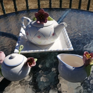 May include: A white tea set with floral accents. The set includes a teapot, creamer, and sugar bowl. Each piece features a delicate floral design with pink and green accents.