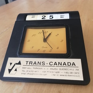 May include: A black desk clock with a gold face and a calendar display. The clock shows the time as 11:55 and the date as Thursday, May 25th. The clock also has the text "TRANS-CANADA" and contact information for the company.