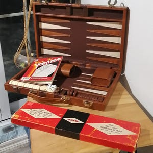 May include: A vintage brown leather case containing a cribbage board. The case is open, revealing the board, dice, pegs, and a rule book for the game Yahtzee. The case is resting on a wooden table. The box for the cribbage board is red with black lettering and reads "Deluxe Travel Size Cribbage Board".