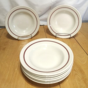 May include: A stack of white ceramic soup bowls with a maroon rim, plus two more on metal stands. The bowls are on a light brown wooden surface, with a white backdrop.