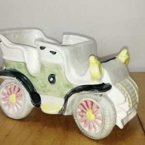 Peut inclure: Un pot de fleurs en céramique en forme de voiture ancienne, dans les tons de blanc, gris, vert et noir. Le véhicule vintage présente un design détaillé avec des accents roses et jaunes sur les roues. Le pot a un dessus ouvert.