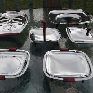 Op de afbeelding: Een set van zes vintage serveerschalen van roestvrij staal. De set bevat ovale en vierkante schalen, een kleine pan met een handvat en een schaal met een hoog handvat. Elk stuk heeft donkerbruine houten handvatten of accenten.