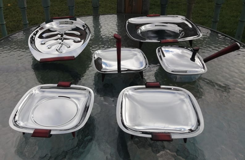 Op de afbeelding: Een set van zes vintage serveerschalen van roestvrij staal. De set bevat ovale en vierkante schalen, een kleine pan met een handvat en een schaal met een hoog handvat. Elk stuk heeft donkerbruine houten handvatten of accenten.