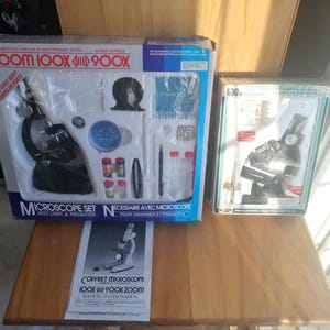 Könnte beinhalten: Zwei Mikroskop-Sets werden auf einer Holzoberfläche präsentiert. Das größere Set, mit der Aufschrift "ZOOM 100X 900X", enthält ein Mikroskop, Objektträger und Zubehör. Ein kleineres Set, mit der Aufschrift "600x", ist ebenfalls sichtbar. Ein Handbuch liegt vor dem größeren Set.
