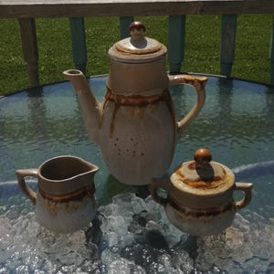 Peut inclure: Un service à café en céramique avec un glaçage beige et marron. L'ensemble comprend une cafetière avec un couvercle, une crémière et un sucrier avec un couvercle. La cafetière a un long bec et une anse. Le sucrier a deux anses.