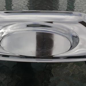 Op de afbeelding: Een rechthoekige serveerschaal van roestvrij staal met een verhoogd midden en houten handvatten. De schaal heeft een gepolijst, reflecterend oppervlak en wordt ondersteund door vier kleine, donkere houten poten. De handvatten zijn roodbruin.