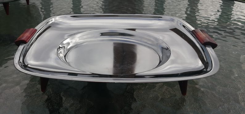 Op de afbeelding: Een rechthoekige serveerschaal van roestvrij staal met een verhoogd midden en houten handvatten. De schaal heeft een gepolijst, reflecterend oppervlak en wordt ondersteund door vier kleine, donkere houten poten. De handvatten zijn roodbruin.