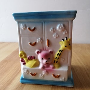 May include: A white ceramic dresser with blue trim, featuring a pink teddy bear, a yellow duck, and a yellow giraffe. The dresser has three drawers with a floral design.