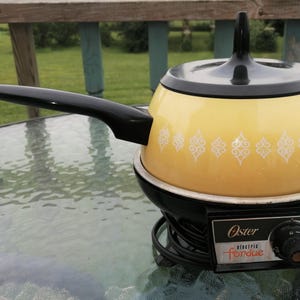 May include: A vintage Oster electric fondue pot. The yellow pot has a black handle and lid. The black base features a temperature control dial and the Oster logo. White decorative patterns are on the pot's exterior.