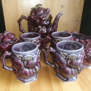 Op de afbeelding: Een set van paarse en gouden keramische theekopjes en een theepot met bloemmotieven. De theepot heeft een deksel en een tuit. De theekopjes hebben handvatten en zijn versierd met bloemen en bladeren.