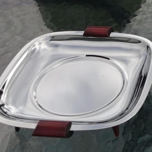 Op de afbeelding: Een glanzende, vierkante zilveren serveerschaal met afgeronde hoeken en een verhoogd cirkelvormig midden. Het heeft donkerrode houten handvatten en kleine pootjes. De schaal is waarschijnlijk bedoeld voor het serveren van voedsel of het tentoonstellen van items.