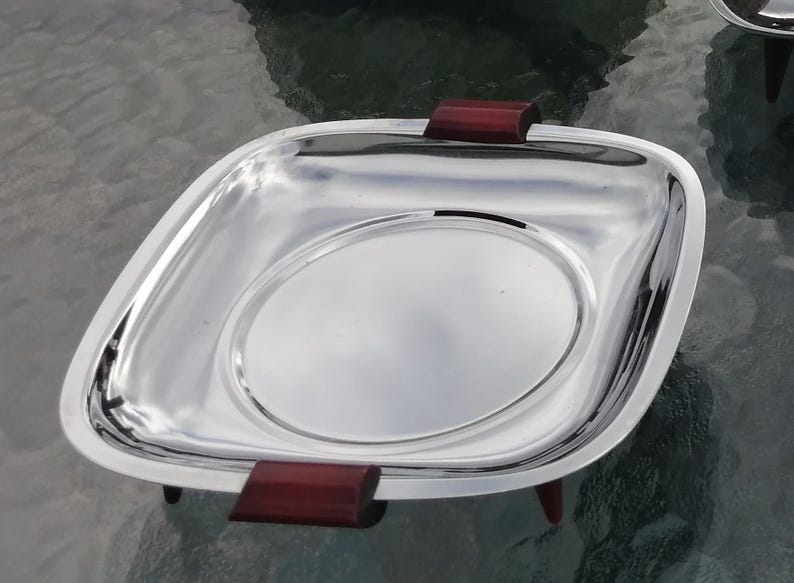 Op de afbeelding: Een glanzende, vierkante zilveren serveerschaal met afgeronde hoeken en een verhoogd cirkelvormig midden. Het heeft donkerrode houten handvatten en kleine pootjes. De schaal is waarschijnlijk bedoeld voor het serveren van voedsel of het tentoonstellen van items.