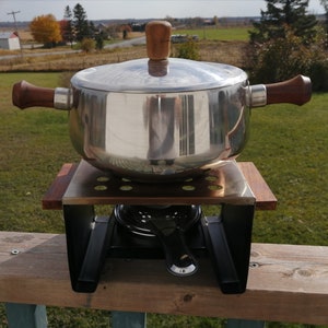 Può includere: Una pentola per fondue d'argento con un manico e un coperchio in legno, appoggiata su un supporto in metallo nero con una base in legno. La pentola è su una superficie in legno.