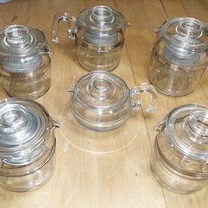 Könnte beinhalten: Sechs durchsichtige Glas-Perkolatoren mit Deckeln. Die Kannen sind übereinander gestapelt und haben einen silbernen Band um die Mitte.