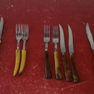 Peut inclure: Une collection de couverts anciens, comprenant des couteaux et des fourchettes, disposés sur une surface rouge. Les couteaux présentent divers modèles de manches, notamment des manches marron foncé, crème et ton bois. Les fourchettes ont des manches jaunes ou marron foncé.