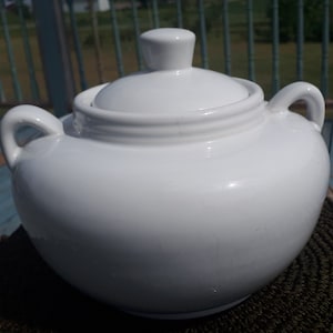 Puede incluir: Azucarero de cerámica blanca con tapa y dos asas. El tazón es redondo y tiene un acabado liso y brillante.