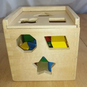 Peut inclure: Un jouet de tri de formes en bois naturel. Le cube présente des formes découpées telles qu'une étoile, un hexagone et un rectangle. À l'intérieur, des formes en bois colorées en rouge, jaune, bleu et vert.