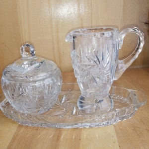 May include: A clear glass sugar bowl and creamer set with a cut glass design. The set is on a clear glass tray.