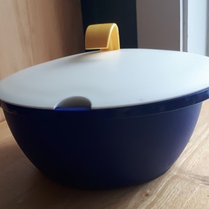 Könnte beinhalten: Eine blaue und weiße Servierschüssel aus Kunststoff mit einem gelben Deckel. Die Schüssel ist rund und hat eine glatte Oberfläche. Der Deckel hat einen Griff oben.