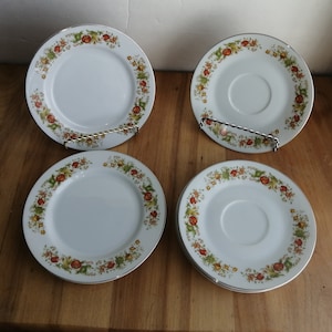 Peut inclure: Ensemble de quatre soucoupes en céramique blanche avec une bordure dorée et un motif floral aux accents rouge, orange et vert.