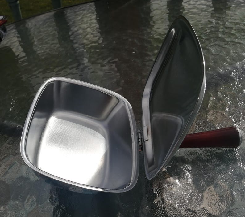 Op de afbeelding: Een roestvrijstalen, vierkante voedselwarmer met een scharnierend deksel. Het deksel is open en onthult de binnenkant. De handgreep is van donker hout. De warmer staat op een glazen tafel.