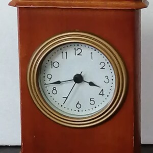 May include: A wooden clock with a gold-rimmed face and black hands. The clock face has black numbers and a white background. The clock is sitting on a wooden base.