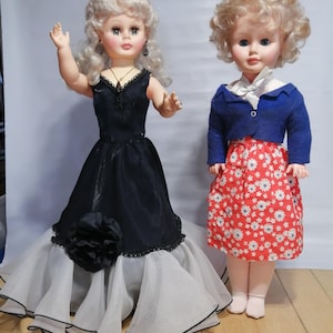 Può includere: Due bambole vintage. Una bambola indossa un abito nero con una gonna bianca e nera e un grande fiore nero. L'altra bambola indossa una giacca blu, una gonna rossa con fiori bianchi e una sciarpa bianca.