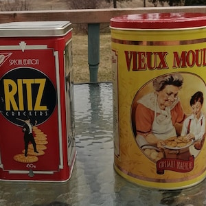 May include: Two vintage metal tins. One is a red rectangular Ritz Crackers tin with a black and yellow logo and a figure. The other is a yellow cylindrical tin with a red lid and the words "Vieux Moulin" and an illustration.