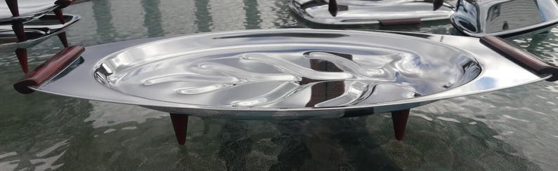Op de afbeelding: Een vintage zilverkleurige serveerschaal met een verhoogd, organisch ontwerp in het midden. De schaal heeft donkere houten handvatten en staat op drie kleine, donkere houten poten. De ovaalvormige schaal is reflecterend.