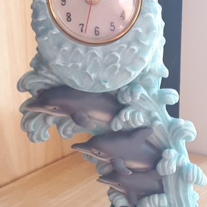May include: A blue and white ceramic clock with a dolphin design. The clock face is white with black numbers and hands. The clock is set against a backdrop of blue waves.