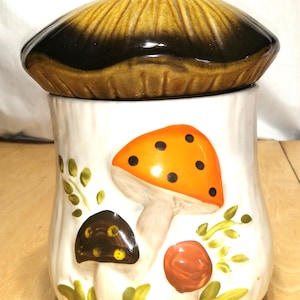 May include: A ceramic mushroom-shaped cookie jar with a lid. The lid is brown and black, topped with an orange ladybug. The jar is white with painted mushrooms in orange, brown, and red, with green leaves.