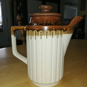 May include: A brown and white ceramic teapot with a ribbed design and a brown lid. The teapot has a brown and white glaze with a speckled finish.