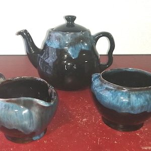 Pode incluir: Um conjunto de bule, jarro de creme e açucareiro de cerâmica preto e azul. O bule tem um corpo arredondado e uma alça lateral. O jarro de creme e o açucareiro têm uma forma arredondada com uma borda na parte superior.