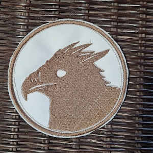 Könnte beinhalten: Runder, gestickter Aufnäher mit einer braunen Adlerkopf-Silhouette auf weißem Grund, umrandet von einem braunen Rand. Der Aufnäher liegt auf einem gewebten, braunen Hintergrund, der einen Kontrast erzeugt.