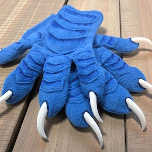 Può includere: Un paio di guanti blu a forma di artiglio di mostro con artigli bianchi. I guanti sono realizzati in un materiale morbido e strutturato e presentano creste dettagliate. Gli artigli sono curvi e appuntiti, aggiungendo all'aspetto realistico.