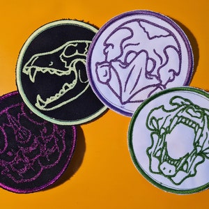 Könnte beinhalten: Vier gestickte Aufnäher mit verschiedenen Designs von Tierschädeln. Die Aufnäher sind rund und haben einen weißen, schwarzen oder hellblauen Hintergrund. Die Schädel sind mit grünem, violettem oder hellgrünem Garn umrandet.