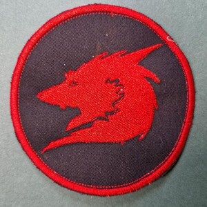 May include: A red embroidered wolf head patch on a black circular background with a red stitched border.