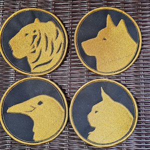 Könnte beinhalten: Vier runde schwarze Stoffpatches mit goldenen gestickten Tiersilhouetten. Die Tiere sind ein Tiger, ein Hund, ein Rabe und eine Katze.