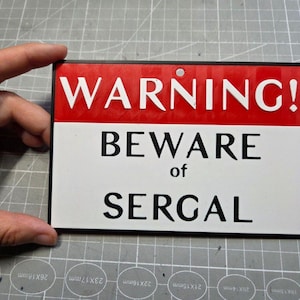 Könnte beinhalten: Rechteckiges Schild mit rotem Oberteil und weißem Unterteil. Schwarzer Text lautet "WARNING! BEWARE of SERGAL". Das Schild hat einen schwarzen Rahmen und ein Loch zum Aufhängen. Maße in Zentimetern.