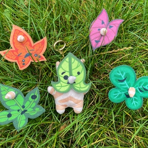 Op de afbeelding: Een verzameling blad-vormige sleutelhangers en een pluche karakter met blad-achtige kenmerken, tegen een achtergrond van groen gras. De sleutelhangers zijn in verschillende kleuren, waaronder oranje, roze en groen, elk met een klein, schelpachtig detail.